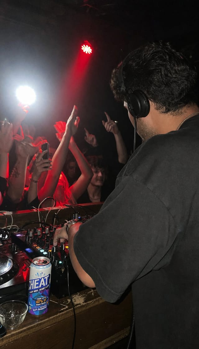 A DJ behind the decks in a small dark club, red light, crowd hands, flash