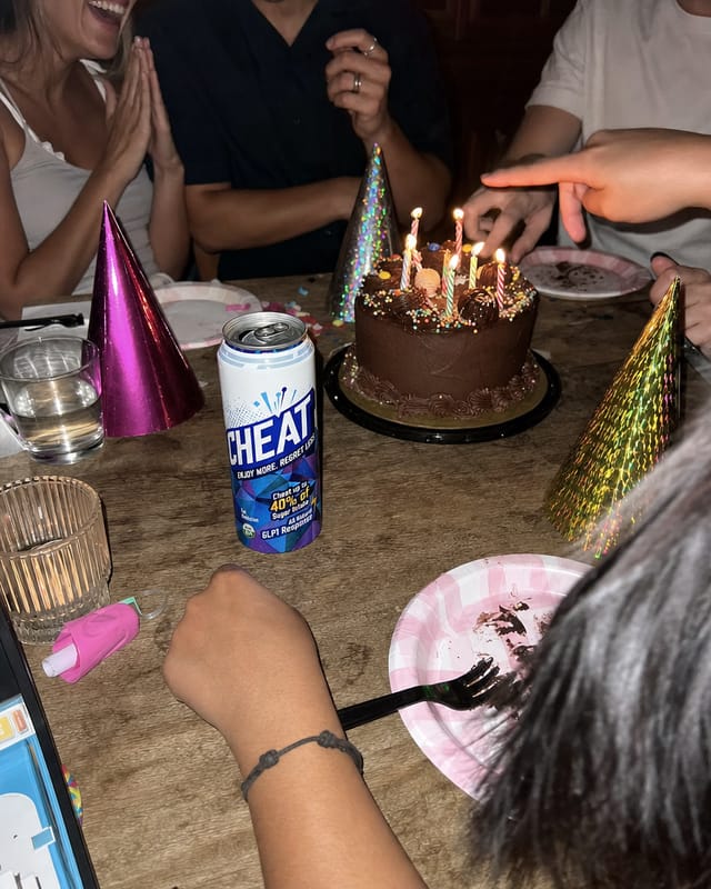 A birthday cake with lit candles on a crowded table, hands, party hats, flash