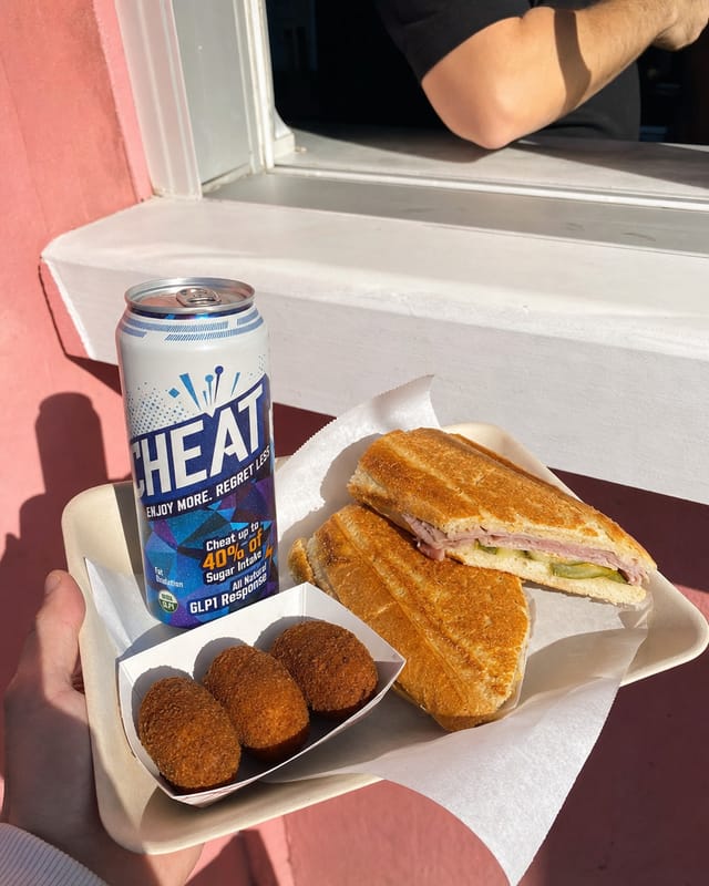 Cuban sandwich and croquetas on a paper tray at a walk-up window, pastel wall, bright sun