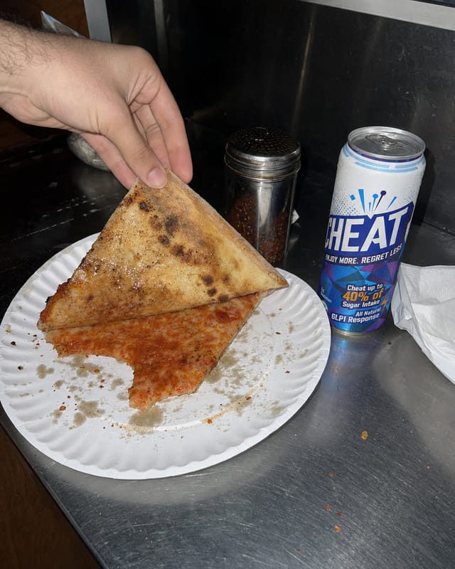 New York pizza slice on a paper plate on a stainless steel counter, grease stains, red pepper shaker, a folded slice being lifted, harsh flu