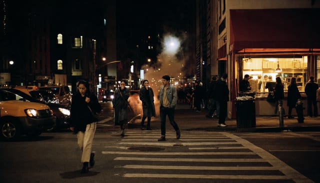 Manhattan street corner at night, pizza counter glowing, steam from a manhole, people crossing, taxis