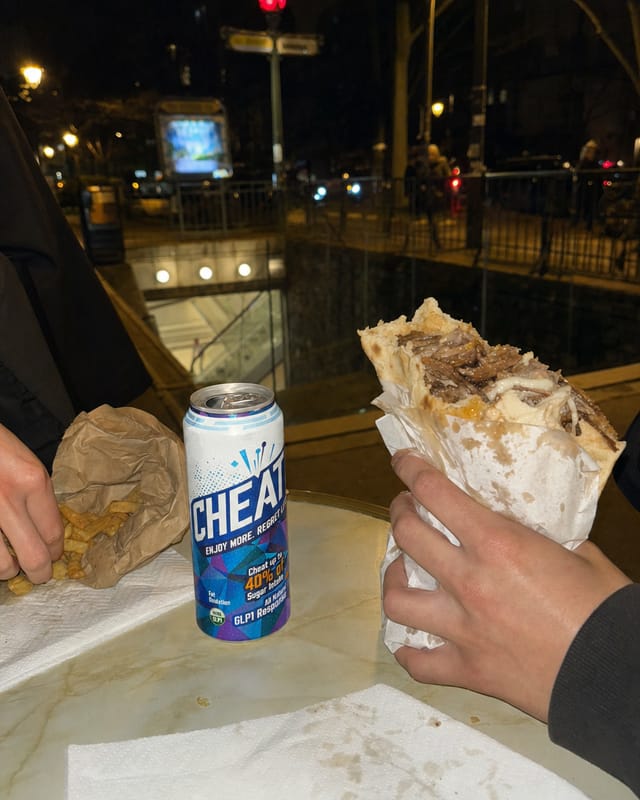 A greasy kebab-frites in paper on a Paris bistro table at 2am, metro entrance visible outside, warm light
