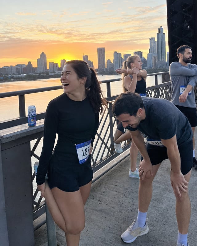 A running club stretching on a bridge at sunrise, race bibs, one runner bent over laughing, city skyline