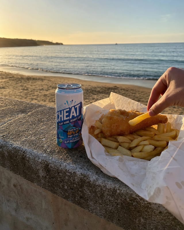 Fish and chips in white paper on a beach wall at golden hour, sand, ocean behind, a hand grabbing a chip