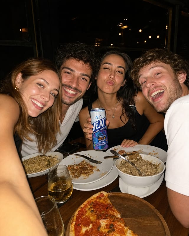 Group selfie at a restaurant table at night, four friends leaning in, flash, plates of food below